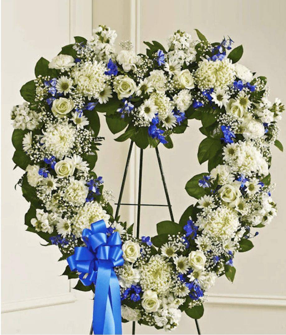Heart-shaped white and blue floral wreath on a stand with a blue ribbon
