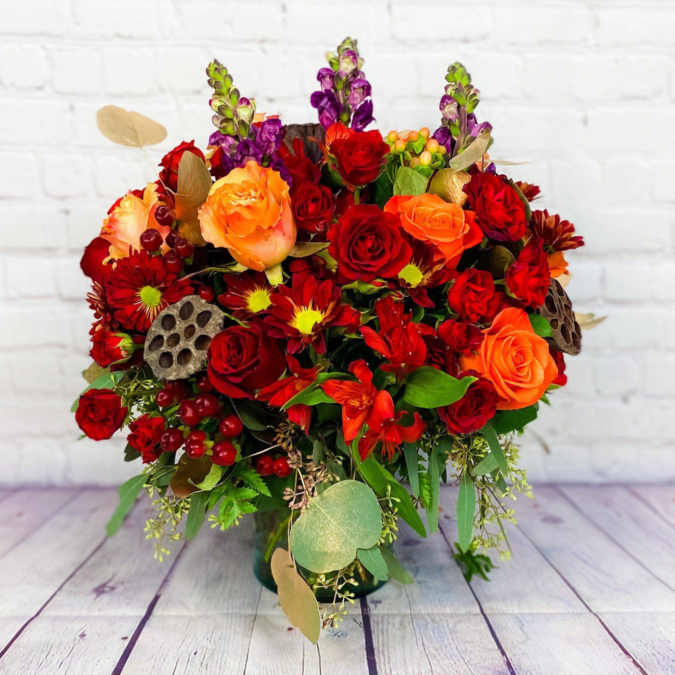Vintage Autumn Blooms by DGM Flowers - Vintage Autumn Blooms is&nbsp;colorful transformation of the autumn landscape transports us to a picturesque countryside from years gone by. Our vintage-inspired arrangement captures that old- world charm with a gathering of blooms in rich, warm tones combined with rustic, natural touches. Handcrafted by our expert florists, it’s a gift that will bring the beauty of the season into the hearts and homes of those you love. •All-around arrangement with red roses, daisy poms and hypericum; orange roses; burgundy carnations and snapdragons; purple waxflower; accented with red painted solidago, seeded eucalyptus, variegated pittosporum, salal, dried oak leaves and lotus pods •Artistically designed inside a clear glass cylinder vase; measures 6"H •Arrangement measures approximately 18"H x 13"L  