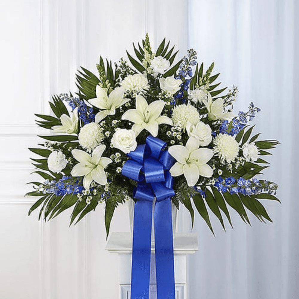 White lilies and blue flowers arranged with a blue ribbon on a stand