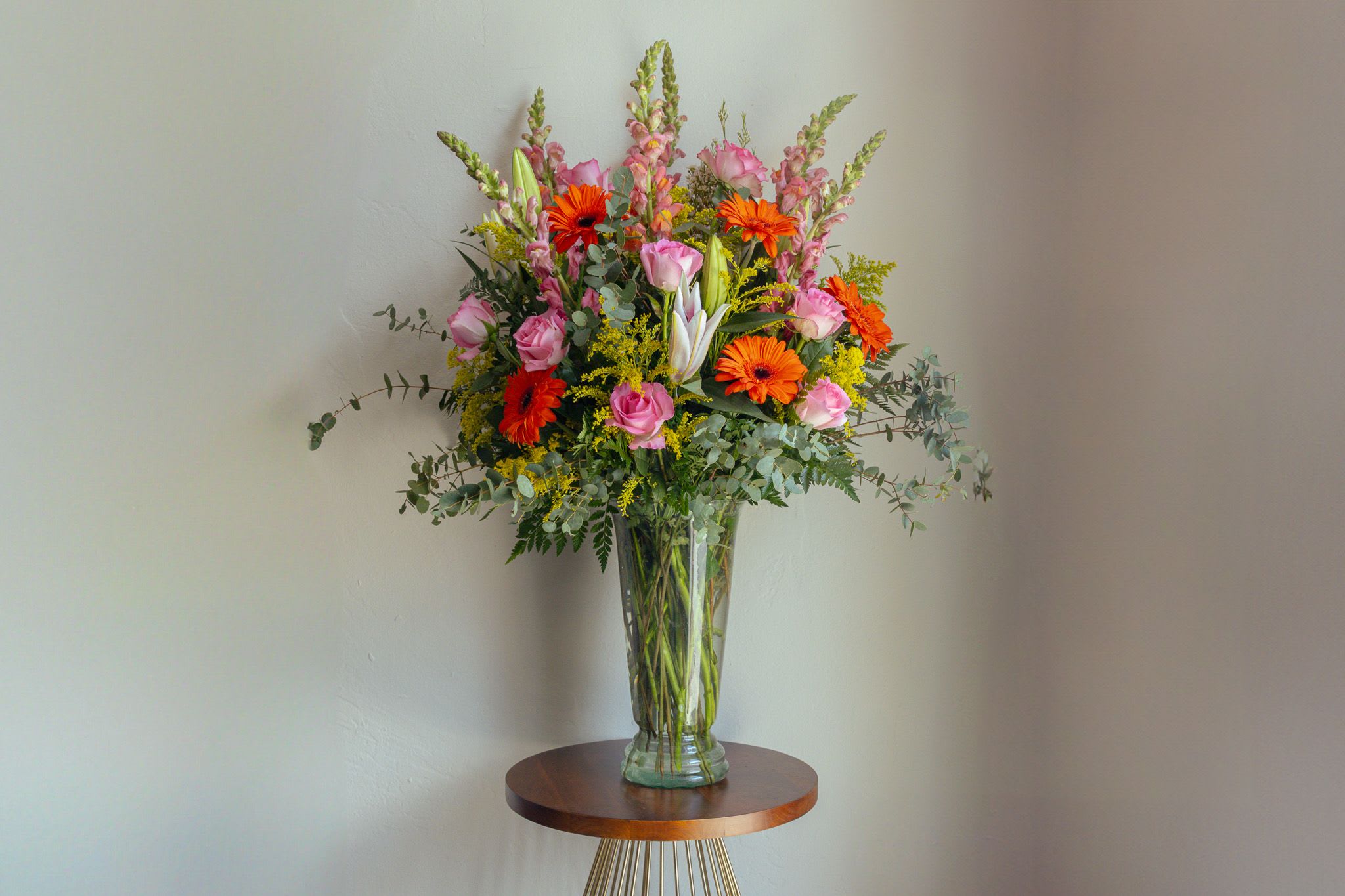 Tall arrangement of pink roses, orange gerbera daisies, lilies and snapdragons in a clear glass vase