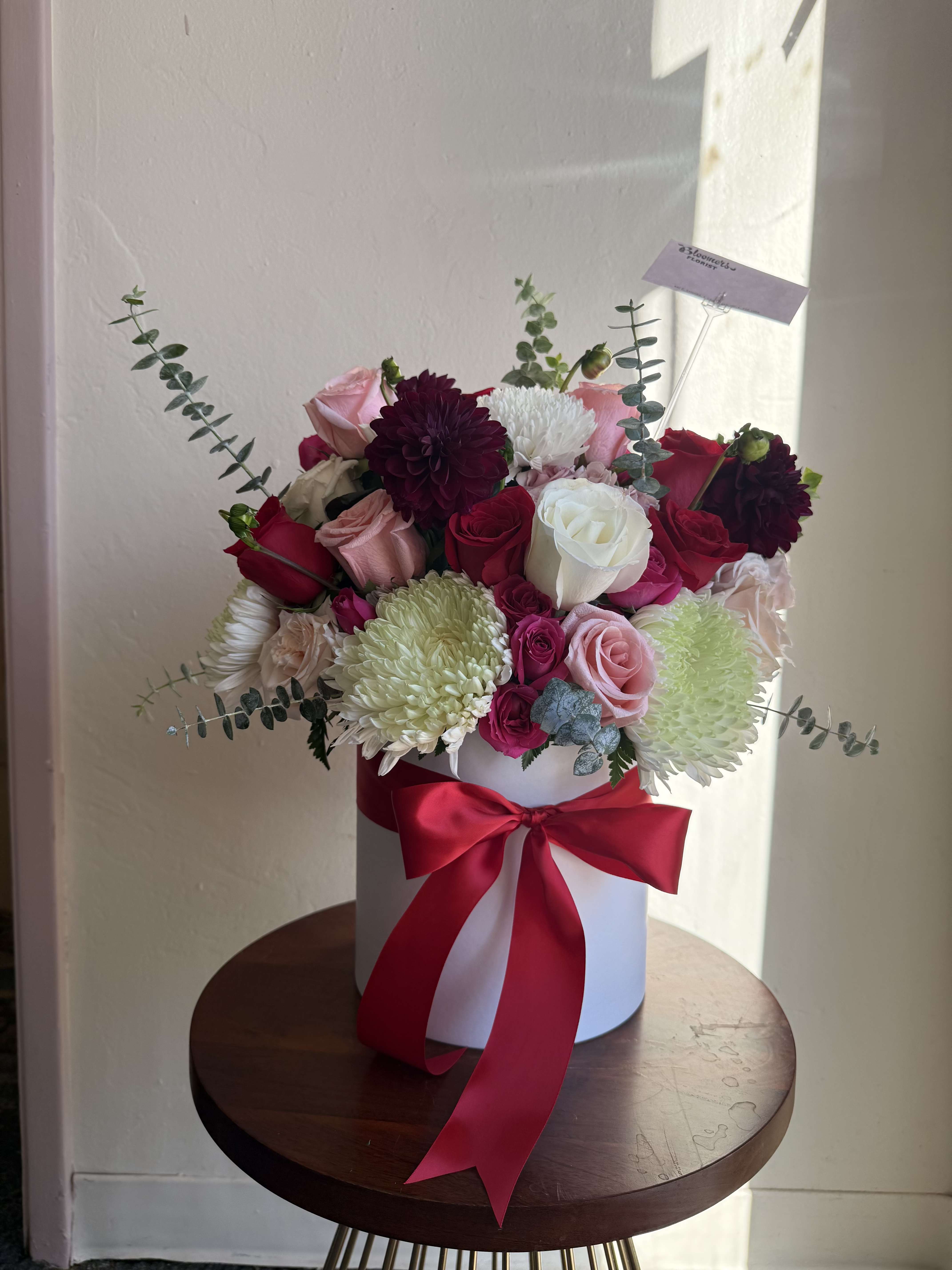 Bouquet of pink, red, white, and burgundy flowers in a white hat box with a red ribbon