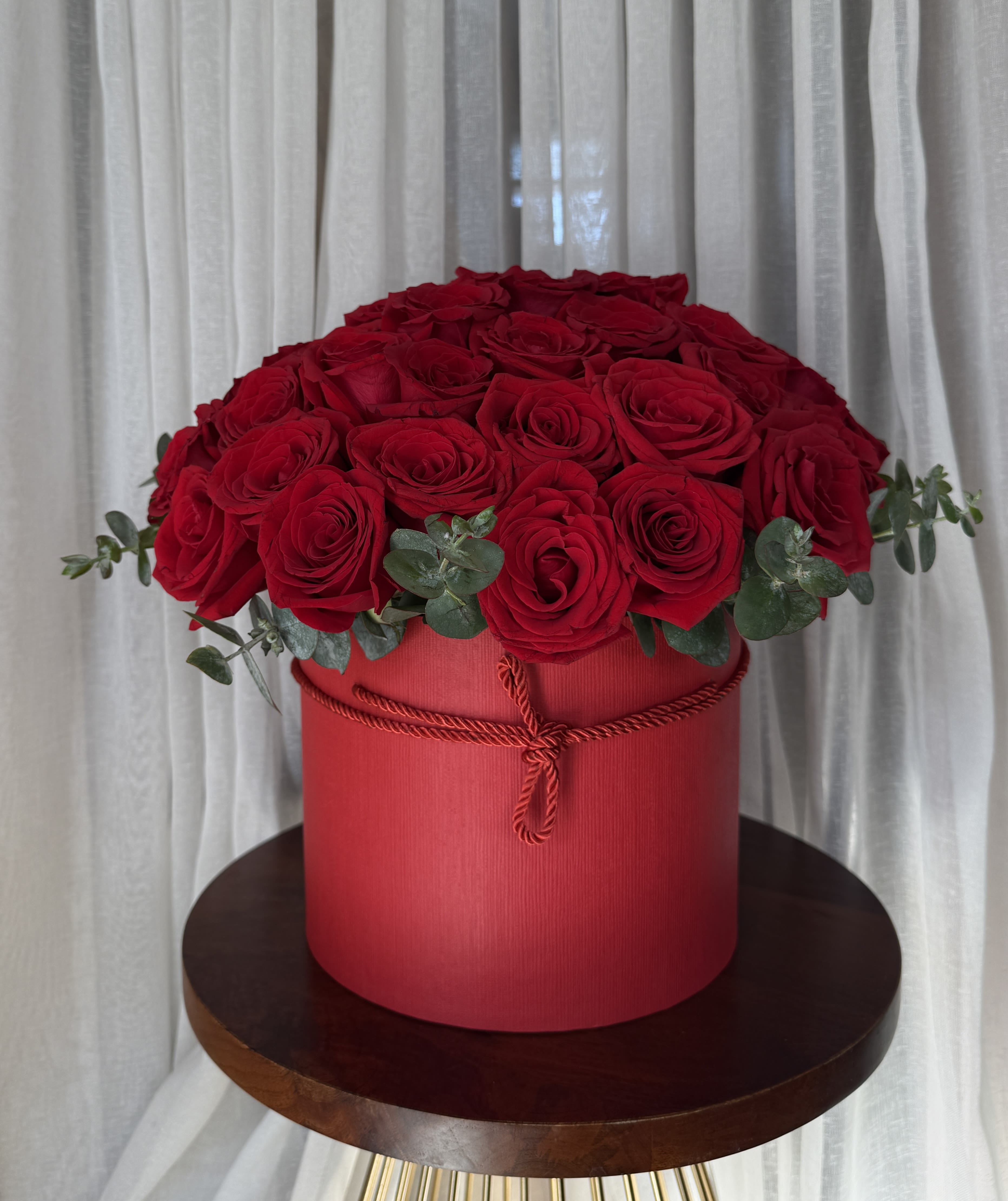 Red roses arranged in a round red box with eucalyptus accents