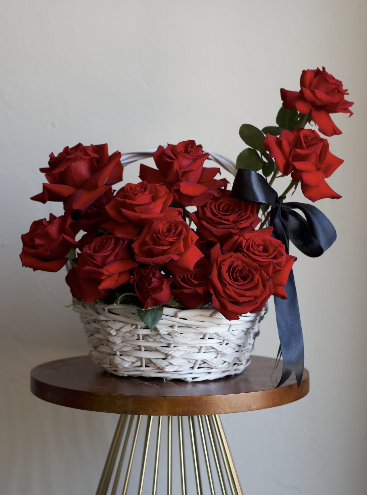 Rose Basket  - A dense cluster of vivid red roses arranged in a white wicker basket creates a timeless romantic statement-the perfect bouquet for an anniversary, Valentine's Day, or any moment you want to make the person you're thinking of feel deeply loved. The rich red petals read as classic and passionate against the clean white basket, while the black satin ribbon adds a polished, formal contrast that makes the arrangement feel intentional and refined.  Additional rose colors are available. Please specify rose color in the notes or give us a call. 