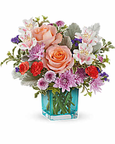 Teleflora's Enchanting Garden Bouquet - Step into enchantment with this delightful turquoise glass cube, showcasing vibrant and colorful blooms inspired by the beauty of a blooming garden. Uncover floral elegance with a garden-inspired bouquet of peach roses, pink alstroemeria, orange carnations, lavender chrysanthemums, purple sinuata statice, and accents of dusty miller and pitta negra. Presented in the Turquoise Cube, this Enchanting Garden Bouquet radiates charm and sophistication.