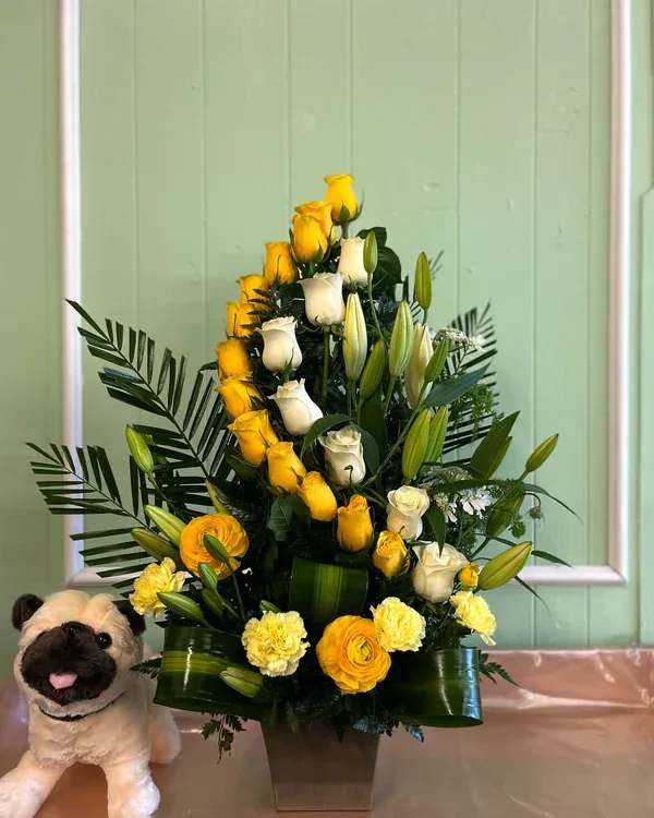 Yellow and white rose arrangement in a square vase with a plush pug beside it