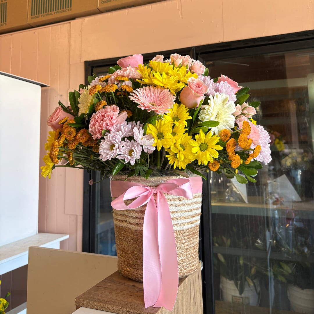 Mixed bouquet of pink, yellow, and white flowers in a woven basket with a pink ribbon