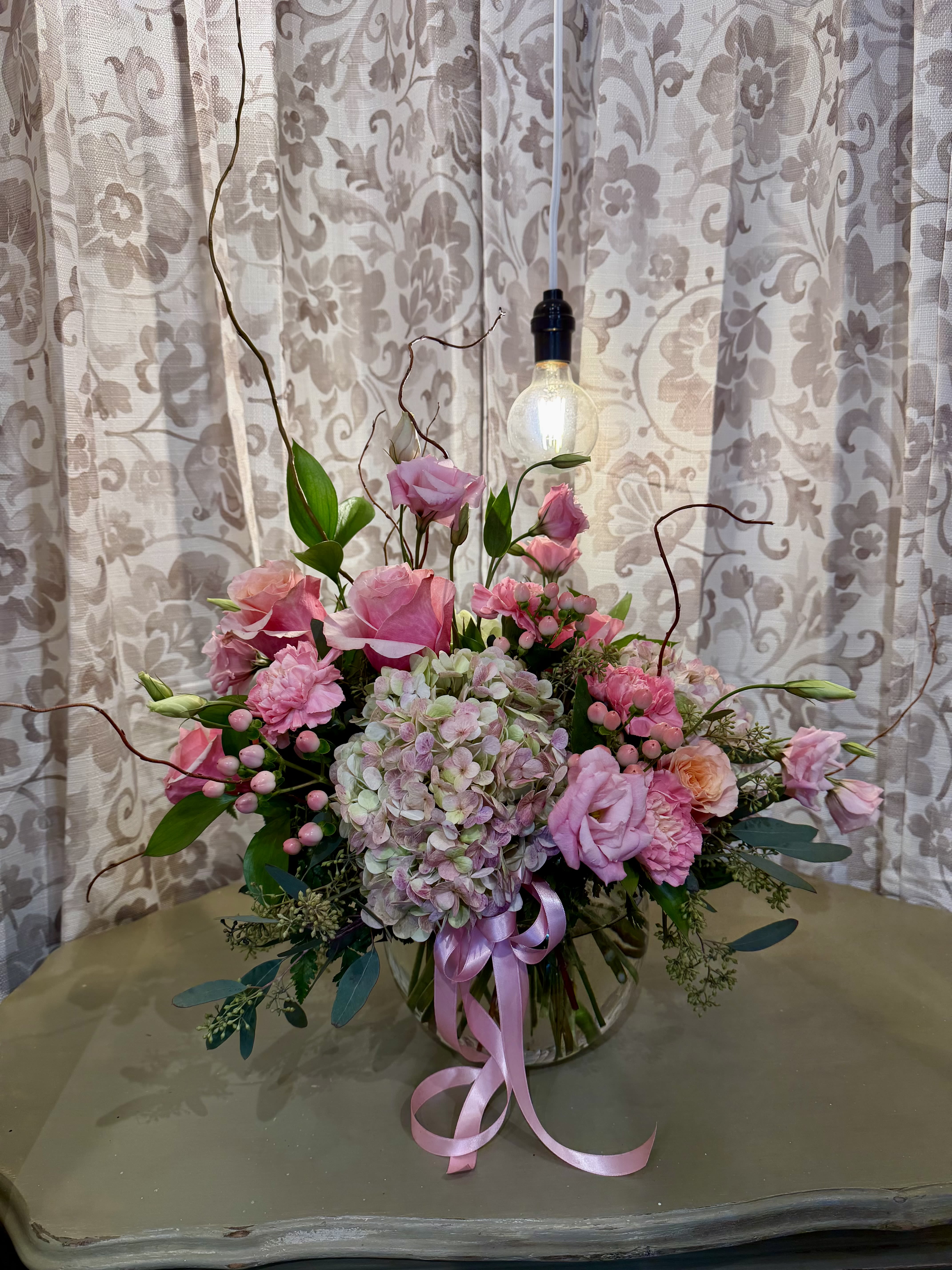 Pink floral arrangement with hydrangeas, roses, and a ribbon in a glass vase