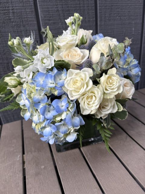 Something Blue   -  Creamy white roses and soft blue hydrangeas form a cool, airy centerpiece that reads calm and elegant - ideal as a winter floral arrangement . The blue-and-ivory palette and rounded, abundant shape make the recipient feel peaceful and seen, whether you're marking a birthday, just because, new arrival to the family, saying thank you, or offering quiet sympathy.  In a  clear square glass vase that shows the clean water line and dense greenery at the base for a fresher, structured feel. Mixed accent blooms and textured foliage nestle around the focal roses and hydrangeas for a full, tactile bouquet that arrives ready to display; place away from direct heat and refresh the water every few days to keep blooms at their best.