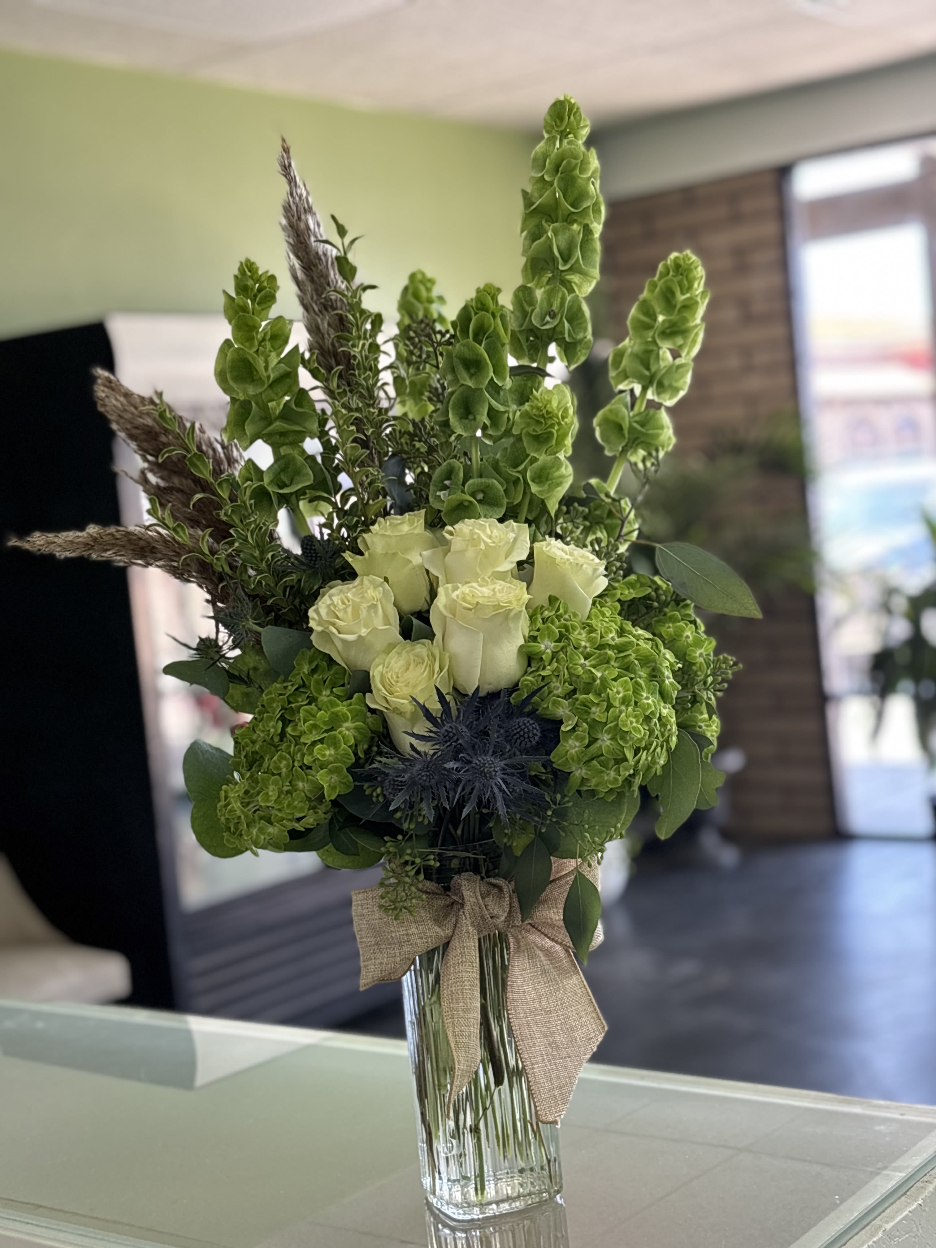 Tall green and white floral arrangement in a glass vase with a burlap bow