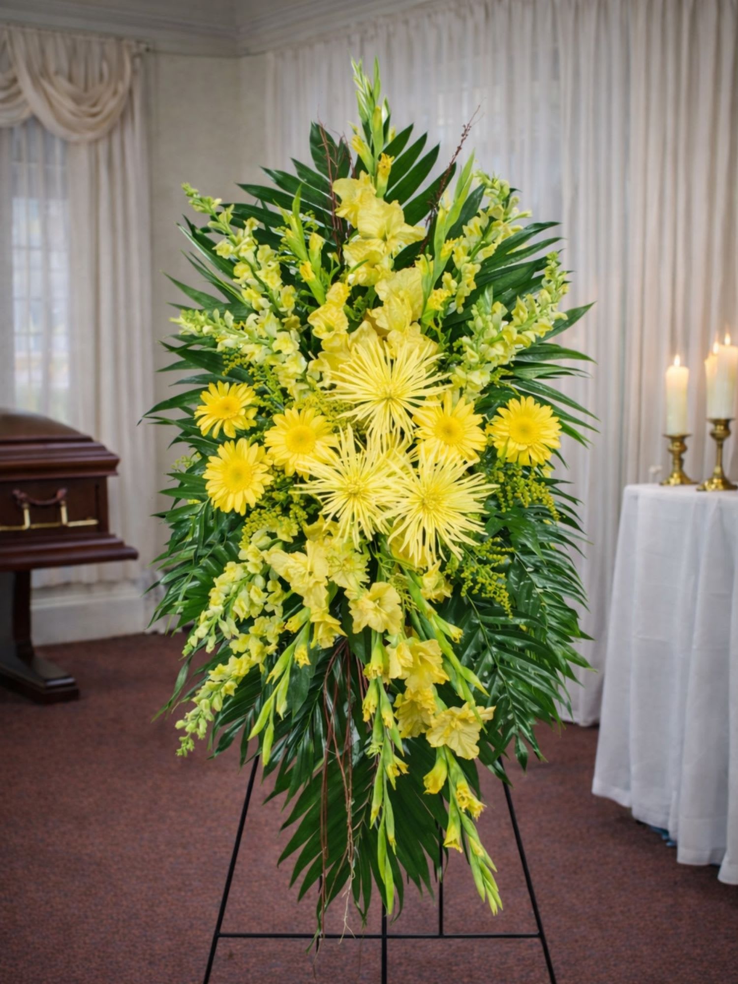Sunny Bright Celebration of Life Funeral Spray - The Sunny Bright Funeral Standing Spray from FloralFabulous is a vibrant and uplifting sympathy tribute created to celebrate the life and memory of someone dearly loved. This radiant arrangement features bold yellow spider chrysanthemums, cheerful gerbera daisies, elegant gladiolus, and delicate snapdragons arranged among lush tropical greenery.  The bright yellow flowers symbolize warmth, remembrance, friendship, and the celebration of a life well lived. Designed as a traditional standing spray, this arrangement creates a beautiful display beside the casket or memorial setting, offering comfort and encouragement to family and friends during a time of loss.  The Sunny Bright Funeral Spray is especially meaningful for celebration of life services where families wish to honor their loved one with flowers that reflect positivity, warmth, and cherished memories.  FloralFabulous proudly provides sympathy flower delivery for funeral services and memorial arrangements in Round Rock, TX and the greater Austin area, helping families express love, remembrance, and support with thoughtfully designed floral tributes.