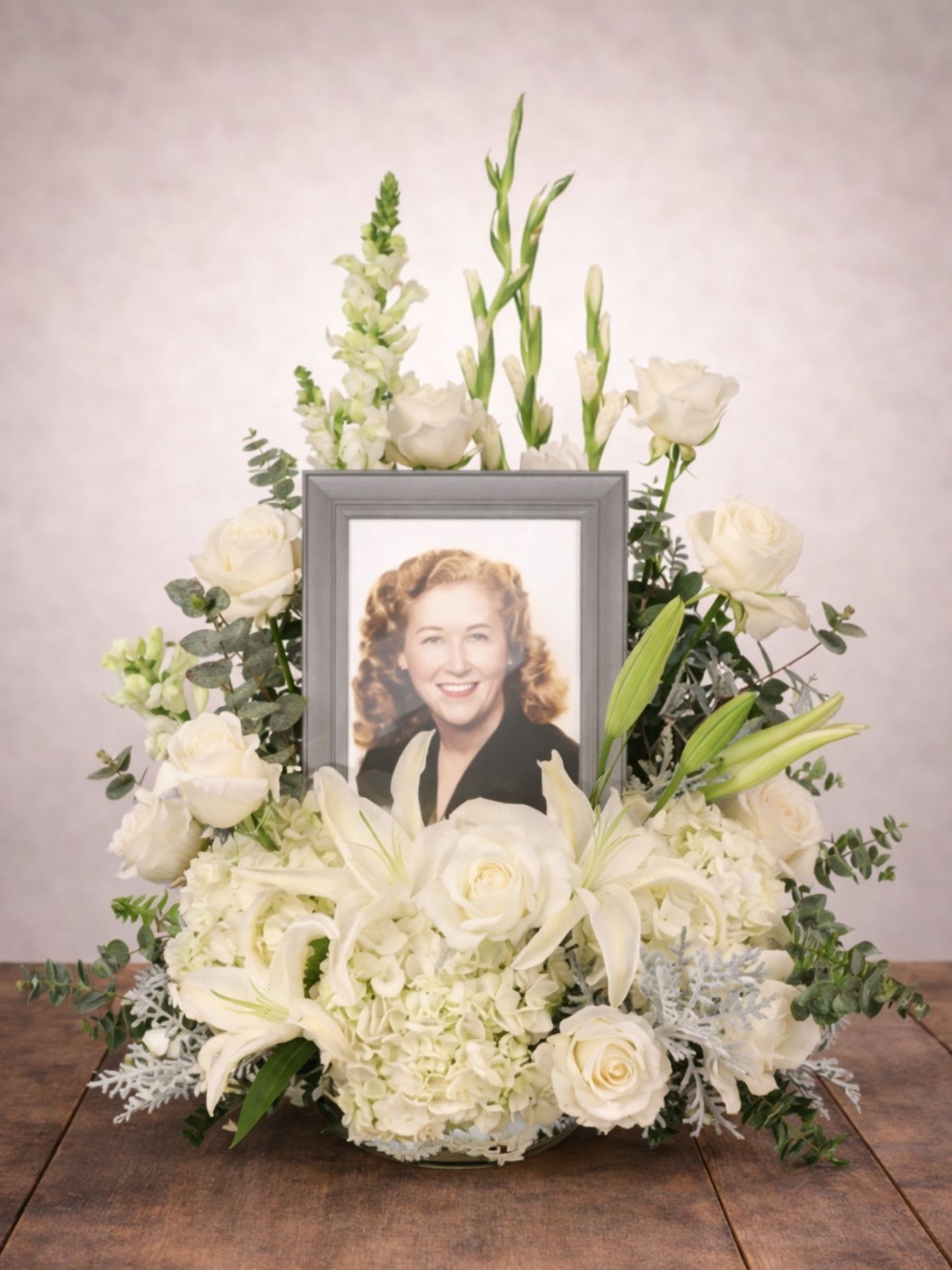 Eternal Peace Tribute Memorial Urn/Photo Arrangement - The Eternal Peace Tribute Memorial Floral Arrangement from FloralFabulous offers a heartfelt and elegant way to honor the life and memory of someone deeply loved. Designed with serene white lilies, classic white roses, lush hydrangea, and delicate snapdragons, this beautiful sympathy arrangement surrounds a framed photograph to create a meaningful tribute filled with love and remembrance.  Accented with soft eucalyptus and silver foliage, the peaceful white palette symbolizes purity, eternal love, and quiet reflection. The framed photo becomes the focal point of the arrangement, allowing family and friends to celebrate cherished memories while surrounded by graceful blooms.  This memorial tribute arrangement is perfect for funeral services, celebration of life ceremonies, and remembrance gatherings. The gentle design provides a comforting presence while expressing heartfelt sympathy and honoring a life well lived.  FloralFabulous proudly offers sympathy flower delivery for memorial arrangements in Round Rock, TX and the greater Austin area, helping families share love, remembrance, and support during life’s most difficult moments..