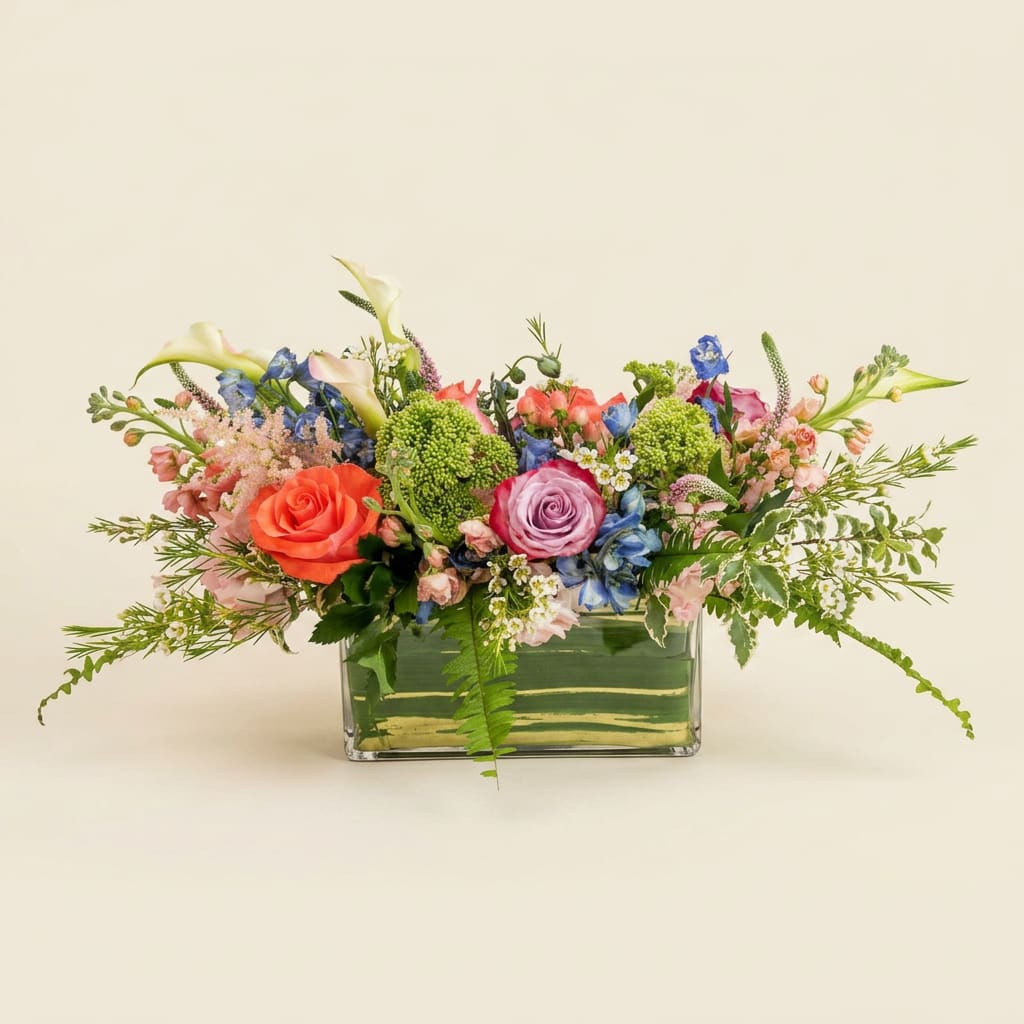 The Glass Garden - Coral and lavender roses take center stage in this garden-style wedding centerpiece, making a joyful first impression for brides and hosts seeking mixed-color floral arrangements for a ceremony or reception. The Glass Garden works beautifully as wedding flowers-its romantic palette of pink, blue, and soft green reads both celebratory and refined, the kind of bouquet that makes the person you're honoring feel genuinely seen on their big day.  Arranged low and wide in a clear rectangular glass vase, the composition pairs roses and calla lilies with small white daisy-like blooms and green button accents for texture and visual movement. Airy stems and trailing greenery spill slightly over the edges, creating a lush, natural silhouette that's polished but not fussy. The mix of bright and pastel tones-coral, lavender, sky blue, crisp white, and verdant green-gives the centerpiece depth and a light, fragrant presence suited to tabletops or sweetheart tables.  Designed for wedding use but adaptable to other celebrations, this arrangement fits nicely down the center of a long table or as an intimate focal piece on guest tables. Its low profile keeps sightlines clear for conversations, and the sturdy rectangular glass vase offers a contemporary, easy-care base-simply refresh the water every couple days to keep blooms looking their best through the event.