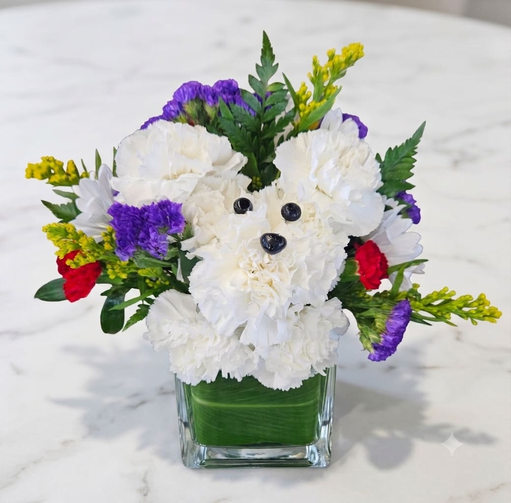 Puppy Love  - Bring a smile to anyone's face with this undeniably adorable, handcrafted floral companion! Perfect for dog lovers, birthdays, get-well-soon wishes, or just because, this unique arrangement transforms fresh blooms into a lovable puppy face that is sure to steal the show.  Features: A sweet puppy face meticulously crafted from crisp, fluffy white carnations, complete with charming eyes and a button nose.  Vibrant Accents: The puppy is nestled in a rich bed of royal purple statice, striking red carnations, and delicate white daisies.  Lush Greenery: Framed with textured leatherleaf ferns and vibrant golden solidago (yellow aster) for a garden-fresh, lively look.  Presentation: Beautifully arranged in a sleek, low-profile green glass vase lined with a fresh leaf wrap for a clean, modern finish.  Perfect For: Birthdays &amp; Celebrations: A fun, memorable surprise for all ages. Get Well Soon: A cheerful pick-me-up to brighten up a hospital room or home. Just Because: Because who could resist those flower-puppy eyes?  Note: Each arrangement is hand-styled by our expert florists using the freshest seasonal blooms, ensuring your puppy pal arrives full of life and personality!