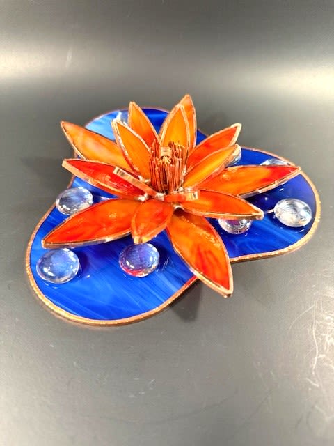 #A002 - Orange Water Lily - A vivid orange stained-glass water lily crowns a marbled blue base in this handcrafted stained glass sculpture - an artful decorative piece perfect as a birthday gift, housewarming accent, or casual celebration present. The striking warm orange and red tones of the layered, pointed petals sit in bold contrast to the deep, swirled blues of the three-lobed base, creating a fiery focal point that draws the eye and brightens any shelf, desk, or tabletop.   The sculpture includes metallic-edged glass petals for crisp definition, a stylized blue stained-glass "pond" base, and several clear glass pebbles arranged around the bloom to catch and reflect light. Finished with a clear glass pebble accent and measuring approximately 2" H x 6 1/2" W x 6 1/2" D, it reads as a modern, small-scale art piece that feels handcrafted and personal. Other colors are available upon request; free shipping within the contiguous U.S. is included, and international shipping is available. 