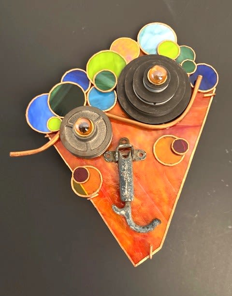 #A093 - "Bubbles" Mask - Bright, whimsical and full of color, this wall-mounted flower arrangement alternative makes a playful gift for birthdays, house warmings, or anyone who appreciates modern, artistic decor. The piece reads as a stylized face-an orange triangular stained-glass base forms the "mask" while layered metal discs and sculpted hardware create bold, expressive eyes and a long, curved nose-so the person you're thinking of will get a laugh and a lasting accent that brightens a wall.   Clusters of round stained-glass circles in blue, green, yellow and multicolored tones rim the top and sides, their smooth glass contrasting with textured rust-like metal for an eclectic, sculptural look. Copper-wire accents, a stained-glass panel, metal hardware pieces and a metal wall-art frame add depth and shine; a metal hook is integrated into the design to suggest a mouth or chin and also makes installation straightforward. This is a compact yet eye-catching decorative piece that sends a lively, artistic message-perfect for someone who loves color, quirky design, or a conversation-starting accent for their home.  Shipping available - select Delivery, then message us to ship.