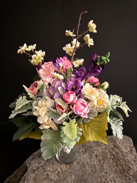 #A051 - Lavender Hydrangea Silk Floral Bouquet - A compact dome of soft pink, cream, lavender and purple silk blooms makes this wedding bouquet a quietly romantic focal point for your ceremony or reception - perfect as a bridal bouquet or a table centerpiece after your special wedding day. The arrangement's mix of rose like blooms, creamy hydrangea clusters and cheerful daisy style flowers is lifted by taller branching sprays of small ivory blossoms to create a natural, garden inspired silhouette that feels both classic and slightly whimsical. Because the flowers are silk, the bouquet keeps its freshly gathered look through photos and celebrations, giving the person you're honoring a lasting keepsake of the day.   Details that matter: large, velvety pale green leaves add texture and rustic contrast to the pastel palette, while deeper purple accents bring depth and visual interest. Options include presentation in a clear glass vase set on a decorative stone slab or as a hand held bouquet wrapped in white satin with delicate pearl beading on the handle for a refined bridal finish. Ideal for weddings, anniversaries, and vow renewals, this silk arrangement arrives ready to display and requires only occasional gentle dusting to maintain its lifelike beauty.  The Lavender Hydrangea, Purple, Pink, &amp; White  Bouquet is a medium sized piece measuring 12" W x 11" D x 14" H.  Shipping available - select Delivery, then message us to ship.