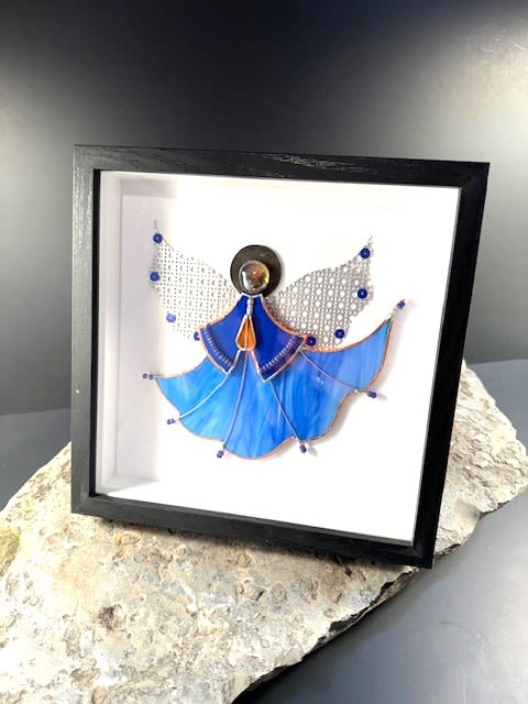 #A061 - Framed Guardian Angel - A luminous stained-glass angel set inside a deep black shadowbox makes an unforgettable sympathy or holiday gift that brings quiet comfort to the person you're thinking of. This framed guardian angel arrangement is ideal for remembrance, Christmas mantel display, or as a thoughtful keepsake for someone needing a gentle reminder they're cared for - they'll feel seen and soothed by its serene presence. The angel's flowing gown is crafted from marbled blue glass outlined in metal, with delicate pale patterned wings and tiny blue bead accents that catch the light. A circular metallic head and a warm amber teardrop at the neckline add contrast to the cool blues, all presented against a clean white backing inside a substantial black shadowbox frame. The piece rests on a rough-textured stone slab base, giving a natural, grounding counterpoint to the handcrafted glass and making the artwork feel both delicate and substantial. Sized to sit on a mantel, shelf, or bedside table, this framed angel is ready to display and requires no special care beyond occasional dusting of the frame and glass. Order it as a comforting memorial keepsake, a meaningful Christmas accent, or a gift to lift someone's spirits-an enduring token that conveys compassion and calm.  Shipping available - select Delivery, then message us to ship.