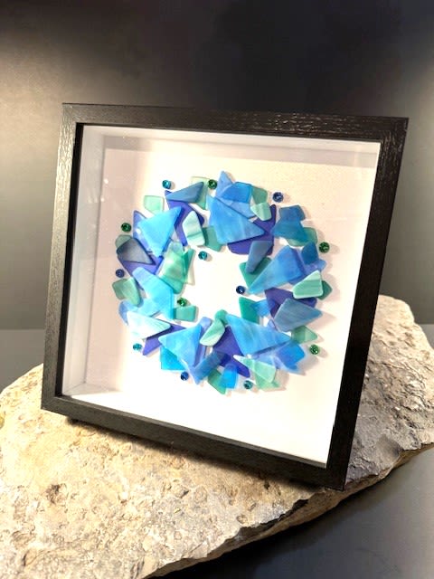 #A082 - Framed Ocean Blue Tumbled Glass Wreath - A coastal-inspired framed wall art piece that reads like a still moment from the shore: layered ocean-blue and teal glass shards form an abstract wreath against a clean white field, set in a sleek black shadow box frame. This floral-style presentation (framed artwork rather than cut blooms) makes a refreshing gift for Mother's Day, Father's Day, housewarming, or any “Just Because” moment when you want the person you're thinking of to feel calm and seen. The composition uses varied sizes and opacities of blue, green, white, and subtle black accents to create depth and movement, with small round glass pieces punctuating the loose circular form. This piece has a contemporary gallery look that suits modern living rooms, offices, or entryways. Ideal for birthdays, retirements, Thank You or I'm Sorry gestures, and seasonal giving like Christmas or Summer décor, it arrives ready to hang and offers a long-lasting, low-care alternative to flowers that will keep evoking a seaside calm for years.  Shipping available - select Delivery, then message us to ship.