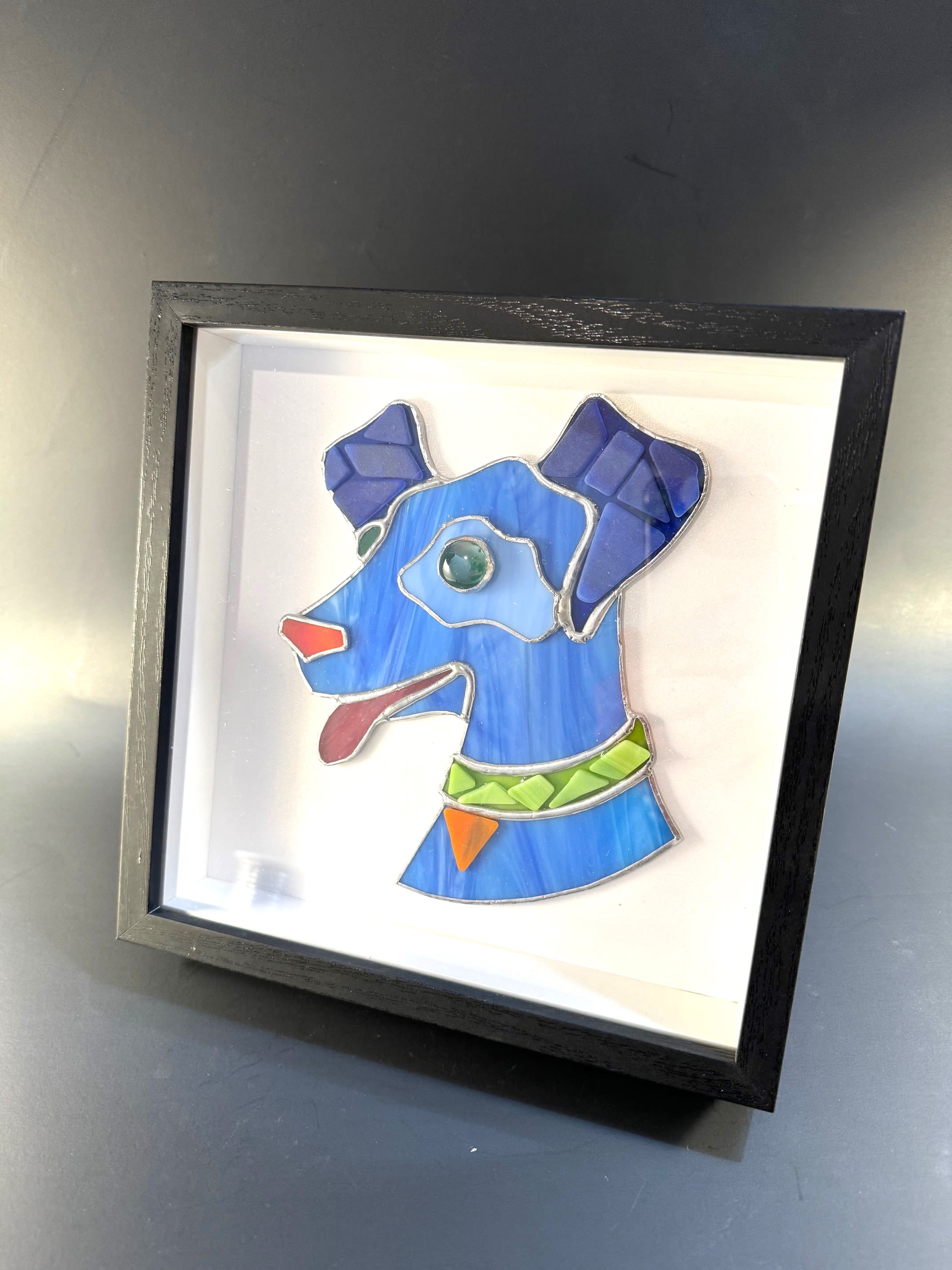 #A032 - Framed Tumbled Glass Dog - A playful stained-glass dog rendered in bright blues, green and orange brings instant cheer-perfect as a framed wall or shelf accent for a birthday bouquet alternative or a Father's Day gift. This framed stained glass art piece works equally well for housewarming, retirement, or "just because" moments when you want the person you're thinking of to smile; pet lovers will feel genuinely seen by the whimsical, modern canine portrait. The artwork features a stylized dog head made of vibrant tumbled-glass-style pieces: primary blues with deeper blue ears and neck, a vivid green collar with orange accents, and a small red tongue for a pop of contrast.   Mounted on a clean white backing and set inside a sleek black shadow-box frame with a glass panel, the piece reads like gallery decor-compact, bold, and easy to place on a wall or shelf to brighten a home or office. Ideal for Christmas stockings, Get Well gestures, Thank You or Thinking of You deliveries, and seasonal touches from spring through summer, this framed dog makes a thoughtful gift for birthdays, Grandparent's Day, Boss's Day, or a cheerful "I'm Sorry."   Shipping available - select Delivery, then message us to ship.