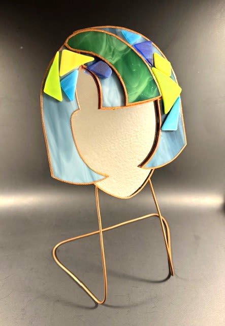 #A117 - Denim Blue Mosaic Face Mirror - A modern stained-glass sculpture that reads like art and affection, perfect as a gift for Mother's Day, a birthday, or “just-because” surprise. This decorative glass face mirror stands firmly on a delicate metal wire stand and brings a bright, playful moment to anniversaries, housewarmings, graduations, or to say "I'm thinking of you." The abstract human silhouette and bold color blocking make the recipient feel intentionally seen and celebrated.   The piece features opaque and translucent glass in soft blues, fresh greens, bright yellow accents and touches of white, each section outlined in warm copper edging to suggest hair and facial contours without literal features. Mounted on a copper wire stand, the stained-glass mirror sits lightly on a tabletop or shelf, adding sculptural height and a splash of color to mantels, desks, or bedside tables. Its clean geometric shapes and cheerful palette work especially well for spring and summer décor but remain striking year-round.   Offer this as a thoughtful option for holidays and milestones - from Christmas and weddings to graduations, retirements, or a thoughtful get-well or thank-you gift. Easy to place and enjoy, just set on a stable surface away from direct impact. The recipient will receive a contemporary keepsake that radiates warmth and personality long after the moment has passed.  Shipping available - select Delivery, then message us to ship.