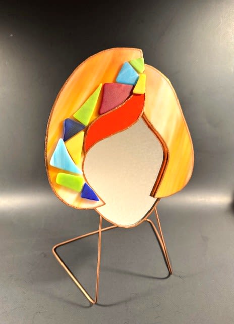 #A059 - Creamsicle Orange Mosaic Face Mirror - A playful, modern stained-glass face sculpture that doubles as a cheerful decorative accent-perfect as a housewarming gift, birthday surprise, or Mother's Day treat. This standing face mirror brings mosaic-like energy to any shelf or desk and helps buyers find the right gift for anniversaries, graduations, or "just because" moments.  Swirled orange glass shapes the head with an open, reflective center, while bold blocks of red, blue, green, and yellow form a geometric accent along one side. The piece is edged in warm copper tones and set on a simple triangular metal stand for a light, airy presence; its compact size and sculptural quality make it an easy fit for contemporary living rooms, home offices, or creative studios. The vivid color mix and abstract lines read lively and artistic-ideal for friends who love color, coworkers for Boss's Day, or someone needing a cheerful get-well surprise.  This is the kind of gift that makes the recipient feel seen and celebrated-whether you're marking Christmas, a retirement, a thank-you, or a moment of romance. Because of its durable glass and metal construction, it needs only occasional dusting to keep the colors bright; choose this piece when you want a long-lasting, decorative reminder that bright days are ahead.  Shipping available - select Delivery, then message us to ship.