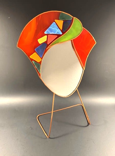 #A050 - Bright Orange Mosaic Face Mirror #1 - A playful burst of color and modern art, this bright orange mosaic face mirror combines bold stained glass with brilliant colors to create a gift that reads as both sculpture and sentiment. Perfect as a Mother's Day, birthday, anniversary, or housewarming gesture, the stylized face-shaped glass with red-orange “hair” and green ribbons catches the eye while the open, lightly frosted center offers a subtle reflective focal point that feels intimate and unexpected for "just because" or thinking-of-you moments. The arrangement pairs vivid red, orange, green, and blue tones-echoing the mosaic's triangular and irregular glass pieces-with complementary blooms and greenery to amplify the sculpture's celebratory mood.   Mounted on a slender metal display stand, the stained glass mirror is mounted on a copper wire stand, making it ideal for a desk, shelf, or side table without overwhelming small spaces. Its compact, modern silhouette and tactile mix of glass and petals make it equally right for graduations, retirements, get-well wishes, or a romantic surprise. Give this to the person you want to delight with color and creativity-the kind of gift that makes them feel seen and celebrated.   Shipping available - select Delivery, then message us to ship.