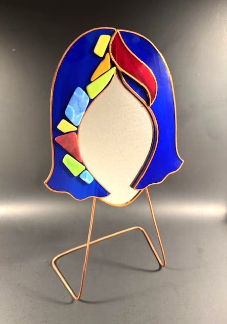 #A053 - Cobalt Blue Mosaic Face Mirror - A cobalt blue stained-glass face-shaped sculpture brings modern art gifting to the forefront - perfect as a Mother's Day gift, anniversary surprise, or a housewarming accent. This stained glass mirror becomes a keepsake given for birthdays, graduations, weddings, or "just because" moments. The recipient will feel seen and celebrated by an object that's playful, colorful, and hand-crafted in spirit.  The piece features a stylized head outline in rich cobalt blue stained-glass "hair" with a mirror "face" that lets you use it to check your makeup, or peek at the hair on the back of your head by angling it toward another mirror. Bold red glass sweeps across the top while lime green, yellow, and light-blue geometric accents add lively contrast along one side; copper-toned leading and a slender metal stand give warm, artisanal support. Sized to sit on a mantel, desk, or bedside table, it functions as a lasting decorative accent for thank-you gifts, get-well wishes, retirements, or to say "I'm sorry" in a thoughtful way-no watering required.  Shipping available - select Delivery, then message us to ship.