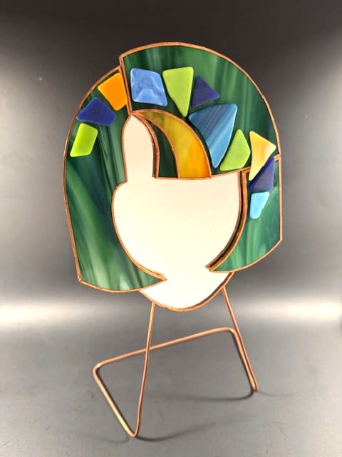 #A076 - Emerald Green Mosaic Face Mirror - A modern stained-glass accent that looks like wearable art - this abstract face mirror is a striking gift for Mother's Day, a birthday, an anniversary, or to brighten a room "just because." The stylized face silhouette of mirrored glass sits at the center, its halo of marbled greens lending a calm, organic feel while pops of blue, lime, yellow, and turquoise add playful light. The overall effect is contemporary and soulful, the kind of piece that makes the person you're thinking of feel seen and celebrated. Crafted with copper-toned metal seams and a simple wire stand, the stained-glass panel is finished in a coordinating metal frame so it stands securely on a desk, shelf, or windowsill. The layered textures-the creamy white center against the marbled green surround and the small geometric color accents-catch sunlight and reflect color into a room without overwhelming a space.   Perfect for housewarming, graduation, retirement, or as a thoughtful get-well or "I'm sorry" gift, this artistic accent suits spring and summer décor as well as holiday displays like Christmas or Hanukkah. Ideal for the recipient who loves design, art, or unique home touches, this piece brings gentle color and personality to everyday corners. The mirror sits on its metal stand for easy placement.  Wipe gently with a soft, dry cloth to keep the glass bright; avoid abrasive cleaners. Shipping available - select Delivery, then message us to ship.