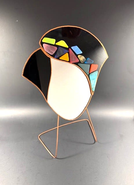 #A040 - Black Mosaic Face Mirror - A striking gallery-style accent that feels like a conversation starter, this abstract stained-glass sculpture makes a bold housewarming or anniversary gift. It is an artistic focal piece-an abstract curved glass panel framed in warm metal and mounted on a slender stand-so it's perfect for Mother's Day, a birthday, or "just because" moments when you want the person you're thinking of to feel truly seen. The design balances dramatic glossy black and opaque white fields with a playful cluster of small geometric glass tiles in yellow, green, blue, purple, and orange. The warm metallic outline and minimalist metal stand add height and a refined contrast to the vivid colors, creating a contemporary, tactile presence ideal for a console, shelf, or bedside table.   Its mixed-color accents let it sit comfortably in modern spring or summer décor and pair naturally with floral gifts for weddings, graduations, retirements, or holiday displays like Christmas and Hanukkah. Give this piece as a thoughtful thank-you, congratulations, or get-well gesture-the recipient will feel celebrated by an object that's both decorative and meaningful. The compact, elevated design fits most tabletops and entryway surfaces; no assembly beyond placing the panel on its stand is required, making it a ready-to-display gift for Mother's Day, housewarmings, or moments when you want to say "I'm sorry," "I love you," or "thinking of you."  Shipping available - select Delivery, then message us to ship.
