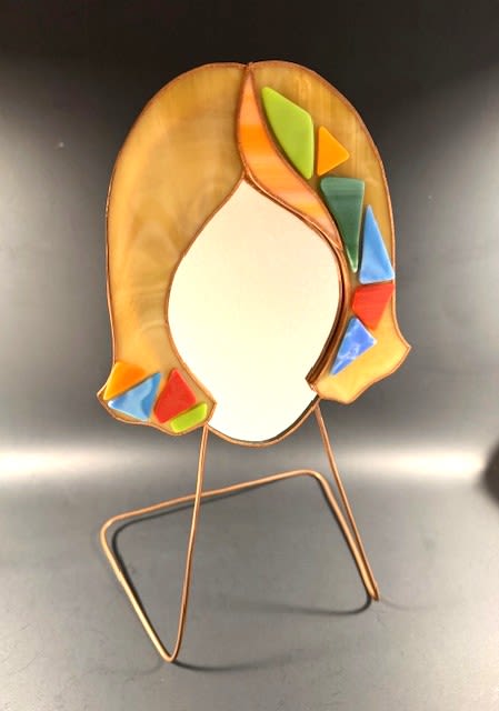 #A010 - Amber Mosaic Face Mirror - A whimsical stained glass face mirror that doubles as artful home décor-perfect as a Mother's Day gift or a thoughtful present for birthdays, anniversaries, housewarmings, or "just because" moments. This sculpture pairs warm amber tones and mixed-color accents with a stylized glass head whose smooth, blank face and sweeping amber hair make an immediate visual statement; the recipient will feel seen and delighted by such a lively, unexpected centerpiece.  The mirror features orange, white, and green tumbled glass pieces arranged in a swish, framed in metal, and set on a simple angular metal display stand. Bright triangular and leaf-like glass inlays-greens, blues, reds, oranges, and yellows-catch light and echo the colors in the rooms decor, adding playful contrast and modern texture. Sized to sit on a mantel, desk, or entry console, the piece reads as both a gift and a long-lasting decorative accent that continues to charm.  Ideal for spring and summer celebrations as well as seasonal holidays like Christmas and Hanukkah, it also makes a memorable choice for graduations, retirements, get-well wishes, weddings, or to say "I'm sorry" or "thank you." Care is simple, display the stained glass sculpture indoors on a stable surface; the metal stand and glass figurine create a keepsake the recipient can reuse as lasting art.  Shipping available - select Delivery, then message us to ship.