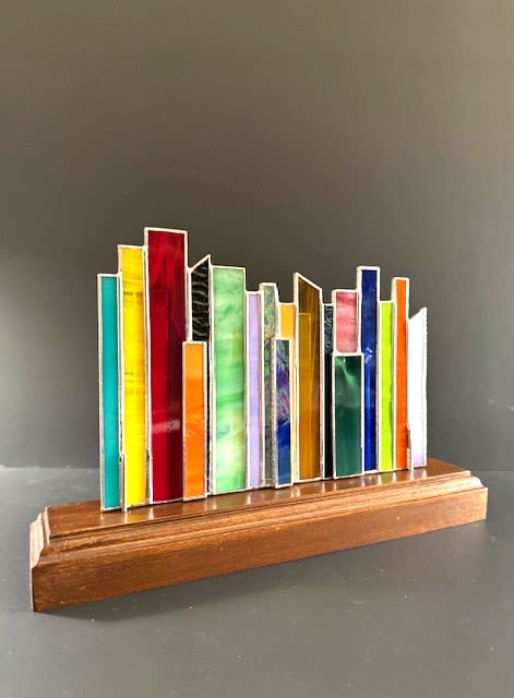 #A018 - Cityscape Standing Sculpture - A vibrant tabletop stained glass sculpture that reads like a miniature city skyline-this mixed-color standing sculpture is an artful gift for birthdays, anniversaries, or Mother's Day. The tall, narrow rectangular panels rise and fall in varying heights and hues-reds, yellows, blues and a full playful spectrum-so the person you're thinking of will see a lively, modern accent that feels celebratory and personal. Its clean, metallic seams give each color block crisp definition, creating movement and light whether placed in bright sun or soft indoor lighting.  Mounted on a polished wooden base, the piece balances warm natural grain with glossy, jewel-toned glass for a contemporary look that fits a desk, shelf, mantel, or bedside table. The sculpture's compact, vertical profile makes it an easy housewarming, graduation, retirement, or get-well gift that doesn't overwhelm a space but still commands attention. Ideal for Father's Day, Christmas, wedding or just-because moments, it's a decorative keepsake that brings color and personality to everyday life. Thoughtful and low-maintenance, this art object needs only occasional dusting to keep its colors sparkling-no watering or special care required-so it's perfect for busy recipients or office display.   Shipping available - select Delivery, then message us to ship.