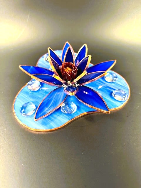 #A056 - Cobalt Blue Water Lily - A sculptural cobalt blue lotus that reads like art and feels like a thoughtful gift - perfect as a decorative art piece for Mother's Day, an anniversary, or just because. The stylized water-lily petals glow in deep cobalt and lighter blue tones of stained glass with warm metallic edging, creating a striking cool and warm contrast that catches light from every angle. This piece makes the person you're thinking of feel seen: calm, celebrated, and surrounded by beauty whether they place it on a desk, windowsill, or bookshelf. Handcrafted glass petals radiate from a textured central core and rest on an irregular, lily pad-like base of marbled light blue glass.   Clear glass droplets and gems dot the base like beads of water, while a complementary stained-glass flower ornament and copper-toned metal accents add layered shimmer and sculptural depth. The compact, luminous design fits easily into home or office spaces and serves as a lasting keepsake for birthdays, housewarmings, graduations, retirements, get-well wishes, thank-you gifts, or holiday presents such as Christmas and Hanukkah. Ideal for anyone who prefers a long-lasting keepsake over fresh blooms, this glass lotus requires no care beyond gentle cleaning.  Shipping available - select Delivery, then message us to ship.