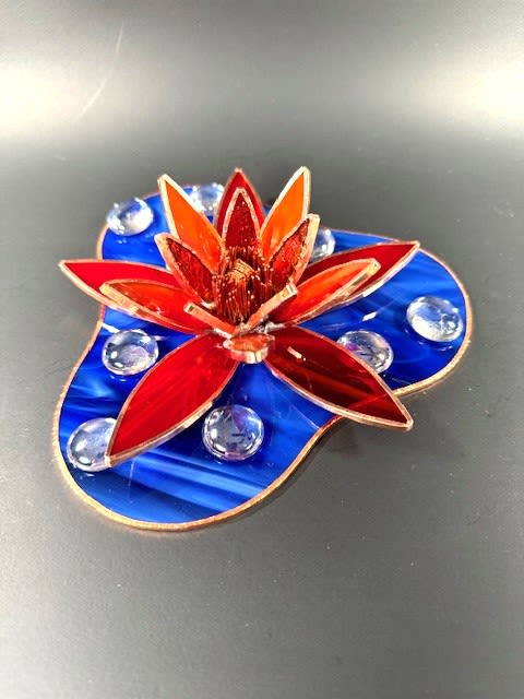 #A108 - Red Water Lily - A bold red and orange stained-glass water lily mounted on a marbled blue base gives this floral arrangement the presence of a small sculpture - perfect as a Mother's Day gift or a standout birthday bouquet alternative. The stylized petals fan outward in layered, pointed shapes that catch light, while clear glass pebbles arranged across the blue background sparkle like water droplets; the result is a vibrant, modern keepsake that reads as both floral art and lasting décor for mantel, desk, or windowsill.   Crafted as a compact decorative arrangement, the piece combines warm metallic edging, a sculpted stained-glass flower figurine, a rounded asymmetrical stained-glass base, and clear glass gems and pebbles for texture and reflection. Its palette of rich red, orange, deep blue, and touches of white creates strong visual contrast-an eye-catching accent for spring or summer but equally at home year-round. Ideal for anniversaries, graduations, house warmings, get-well wishes, or "just because" moments, it's the kind of gift that lets the person you're thinking of feel seen; order as a thoughtful thank-you, wedding favor, or retirement keepsake that doesn't need watering and arrives ready to display.  Shipping available - select Delivery, then message us to ship.