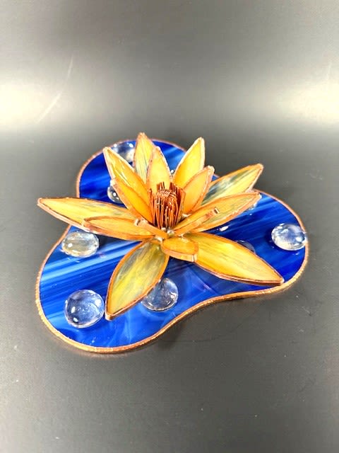 #A128 - Yellow Water Lily - A sunlit, sculptural flower in warm yellow and orange stained glass catches the eye and the heart-perfect as a Mother's Day or birthday gift that feels both artistic and intimate. This flower arrangement-style decorative piece reads like a tiny tableau: layered, pointed petals radiate from a textured center, the copper-colored edging giving each petal a defined, handcrafted warmth that makes the recipient feel truly seen.  The bloom rests on an irregular blue stained-glass base that suggests rippling water, with clear glass gems scattered like dew or pebbles to catch and reflect light. The color palette-cheerful yellow and orange petals against deep blue-keeps the look fresh for spring and summer while working beautifully for housewarming, anniversary, thank-you, graduation, retirement, or just-because moments. Sized for tabletop display, it arrives ready to place on a mantel, bedside table, or office desk and requires no maintenance-an enduring keepsake that brightens a room and the day of the person you're celebrating.  Shipping available - select Delivery, then message us to ship.