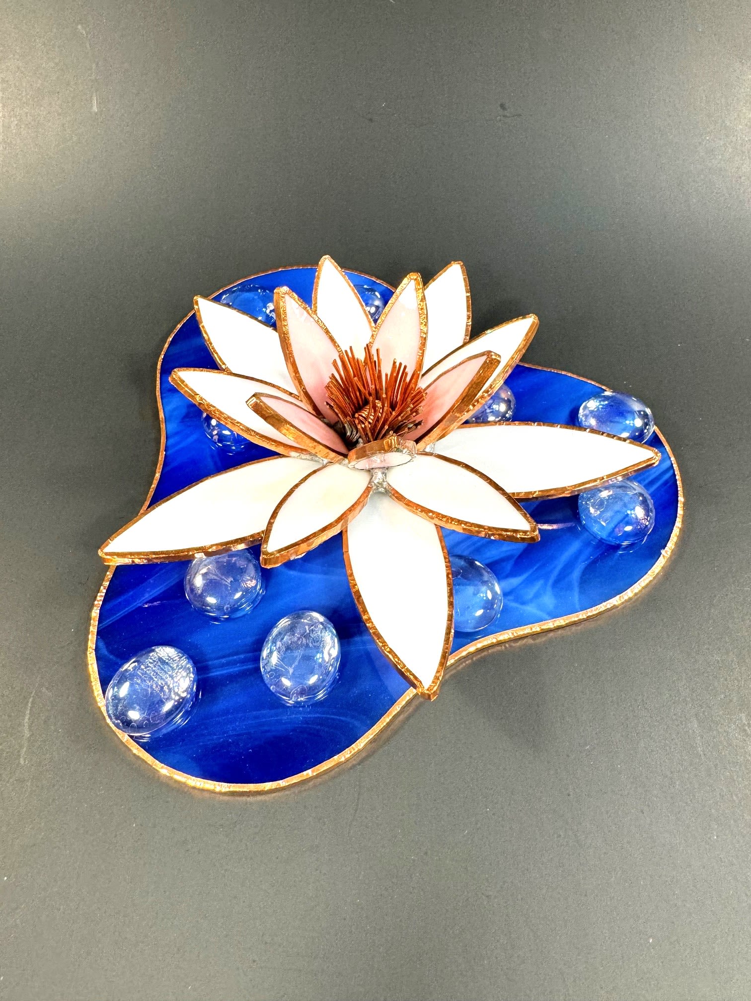 #A134 - White Water Lily - A layered stained-glass waterlily in soft white petals with a blush-pink core, edged in warm metallic trim-this decorative sculpture doubles as a keepsake gift and an artful accent for Mother's Day, anniversaries, birthdays, or "thinking of you" moments. The sculpted cobalt-blue glass base reads like a tiny pond, and the composition's serene palette of blue, white, and pink gives the recipient a quiet, reflective impression that suits gifts for Mother's Day, Get Well, Graduation, or just-because surprises.   Handcrafted details include a stained-glass flower rising from a clover- or heart-shaped deep blue glass base, accented with clear glass pebbles that catch light like floating droplets. The piece works beautifully on a desk, shelf, or bedside table as a small tabletop accent or wedding keepsake; it brings the tactile warmth of handcrafted glass without care-intensive upkeep. For housewarmings, retirements, Father's Day, or as a thoughtful "I'm sorry" or "thank you," this sculpture offers a lasting, elegant reminder that someone is being genuinely seen.