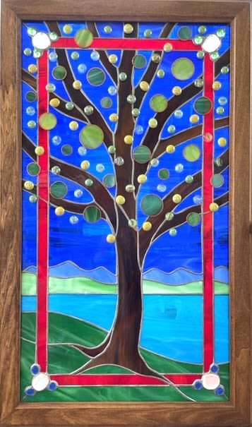 #A048 - Summer At The Lake - Stained Glass Window - Our “Summer At The Lake” panel that feels like a summer afternoon by the water - perfect as a housewarming gift, anniversary surprise, or Mother's Day present. This rectangular stained-glass window captures a stylized tree against a vivid blue sky and shimmering lake, blending artful color and light to make the person you're thinking of feel seen and celebrated. Its vibrant blues and greens with pops of red and yellow give a joyful, window-like focal point that suits birthdays, graduations, or a "just because" moment. Crafted with layered blue and green glass suggesting rolling hills and a reflective lake, the dark brown tree trunk and branching limbs rise inside a striking red border. Round glass accents in green, yellow, and clear tones dot the branches like abstract fruit or lights, while softer blue and green bands hint at distant mountains and horizon.   The panel is set in a warm wood frame that lends a handcrafted, natural finish and makes it ready to hang as wall art - an ideal decorative choice for weddings, retirements, thank-you gifts, or thoughtful apologies. Give a gift that doubles as lasting decor: this piece brightens entryways, sunrooms, or above mantels and carries seasonal charm through spring and summer into the holidays. It arrives framed and ready to display; recommend hanging where morning or afternoon light will catch the glass to bring out the colors and texture, ensuring the recipient enjoys its calming, uplifting presence for years.