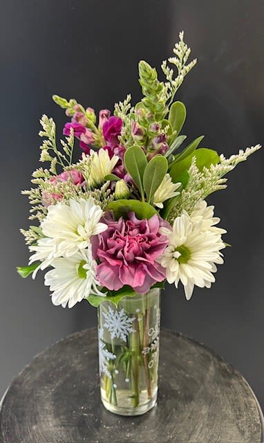 #A077 - Snow Day! - A crisp combination of white chrysanthemums, mauve carnations, and magenta snapdragons creates a fresh, wintry bouquet that feels like a gentle snowfall in a vase - perfect as a winter birthday bouquet, an anniversary surprise, or a get-well arrangement. The upright, compact shape and soft purple-pink palette also make this a thoughtful "thinking of you," "I'm sorry," or "just because" gift that reads as both cheerful and calming for the person you care about. Clustered in a tall, narrow clear glass cylinder printed with white snowflakes, the arrangement pairs daisy-style white mums with ruffled mauve carnations, vibrant snapdragons, and delicate filler blooms for texture and movement. The snowflake vase adds seasonal charm for Christmas or New Year while keeping the look versatile for year-round occasions like Valentine's Day, thank yous, or small desk-centerpiece use. Sized to fit a bedside table or office desk, it's an easy-care choice that brightens a room and makes the recipient feel genuinely seen.