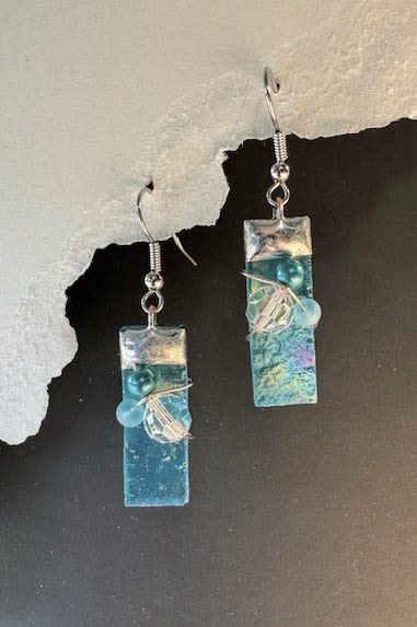 #A046 - Aqua Iridized Stained Glass Earrings - Aqua-tinted rectangular drops catch the light with ocean-blue shimmer-the kind of jewelry that makes the person you're thinking of feel noticed. This flower-shop listing presents a pair of dangle earrings styled like artisanal blooms in blue, black, and white tones, perfect as a Mother's Day gift or for anniversaries, birthdays, and "just because" moments when you want to brighten someone's day.  Each earring features an elongated turquoise-aqua glass rectangle topped with a metallic silver cap and finished on a simple silver-tone hook for a modern, wearable silhouette. Small round and faceted beads are wire‑applied to the glass face, adding delicate texture and sparkle that reads like dewy petals against cool, glassy surfaces. The palette and clean lines suit spring and summer wardrobes but translate beautifully into winter holiday outfits or prom and wedding looks.  Give these earrings to a woman celebrating a graduation, saying "I'm sorry," thanking a friend, or marking a romantic milestone-she'll feel genuinely seen every time she wears them. Lightweight and easy to pair, they arrive ready to gift for Christmas, Valentine's Day, Mother's Day, or simply to say "thinking of you."  Shipping available - select Delivery, then message us to ship.