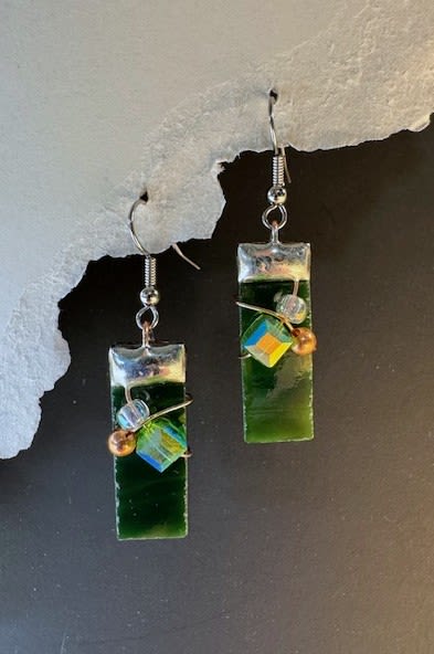 #A081 - Deep Green Stained Glass Earrings - Deep, jewel-toned green glass catches the light in these artisan dangle earrings - an artful accessory that reads like a wearable stained-glass window. Perfect as a birthday gift, Mother's Day surprise, anniversary token, or a "just because" pick-me-up, these earrings give the person you're thinking of a polished yet organic pop of color that works from casual daytime to holiday parties. Each handcrafted pair features long rectangular deep-green glass drops topped with a sleek silvery metal cap and hook. A delicate wrapped-wire accent holds tiny iridescent beads-a small cube with blue-and-yellow shimmer and a round metallic bead – each of which add subtle contrast and movement. The metal findings keep the silhouette modern and light enough for everyday wear while retaining an artisanal, textured feel. Give these for graduations, retirements, weddings, or to say "I'm sorry" or "thank you"-they suit spring and summer wardrobes as well as autumn and winter layers. Lightweight and easy to wear, they arrive ready to gift and make a thoughtful present for a friend, partner, or mother who appreciates handmade, contemporary jewelry.  Shipping available - select Delivery, then message us to ship.