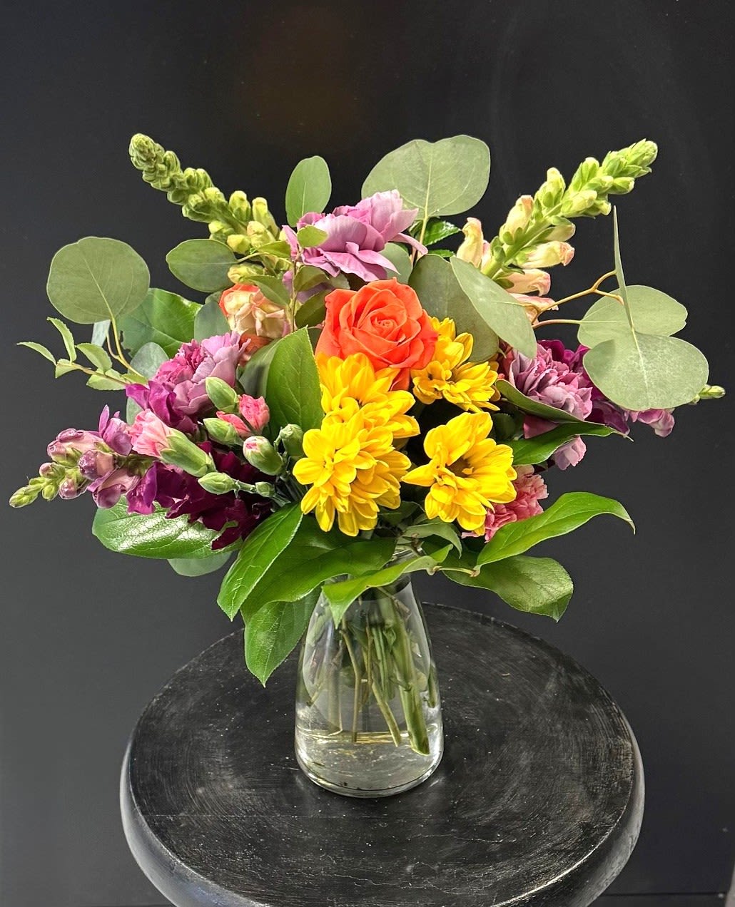 #A112 - Pop of Sunshine - A cheerful burst of color greets the recipient with this bright mixed flower arrangement-a joyful flower arrangement perfect for birthdays, Mother's Day, get well wishes, or “just because”. Yellow chrysanthemums cluster at the front for an immediate sunny impression, while pink and deep purple blooms weave through the design to add depth and warmth. An orange rose near the center serves as a warm focal point, and tall snapdragon stems reach upward to create movement and a lifted silhouette that reads as celebratory and thoughtful.   Carefully gathered in a simple clear glass vase that shows the stems and water, the mix includes carnations, daisies, roses, snapdragons, and chrysanthemums in a vibrant pink, purple, yellow, and orange palette. The casual, approachable styling makes it right for a wide range of occasions-anniversary, graduation, housewarming, new baby, retirement, sympathy moments like Memorial Day, or as a romantic accent for Valentine's Day-while still feeling seasonally appropriate for spring and summer. The visible stems keep the look fresh and easy to reposition in the home; recommend changing the water every two days and trimming stems slightly to prolong bloom life.