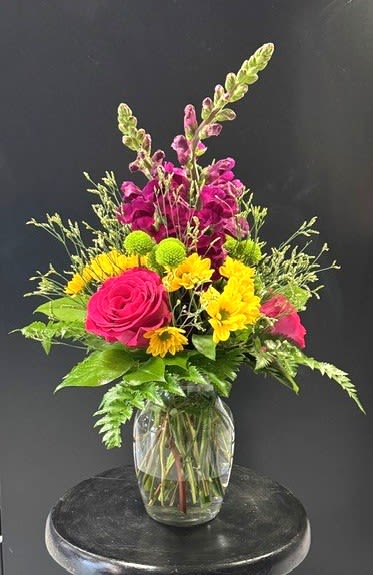 #A013 - Happy Birthday! - Hot pink roses and tall magenta snapdragons create an energetic vertical silhouette in this birthday flower arrangement, a joyful pick for celebrating someone special. The upright shape and saturated palette of red, pink, yellow, and green read as exuberant and fun-perfect for a birthday, congratulations, or simply to brighten a room so the recipient feels noticed and celebrated.  This bouquet pairs hot pink roses with vivid yellow daisy-style chrysanthemums, bright green button mums, and airy filler stems for texture and movement. The snapdragons add height and a playful, architectural line while clustered blooms form a cheerful central focal point. All stems are gathered in a rounded clear glass vase that shows the bundled stems in water for a fresh, casual presentation approximately suited for tabletop display.  Send this when you want someone to feel genuinely seen-whether it's a milestone birthday, a pick-me-up, or a thank-you moment. Low-maintenance care: change the water every 2-3 days, trim the stems at an angle when refreshing the vase, and keep away from direct heat to extend the display.