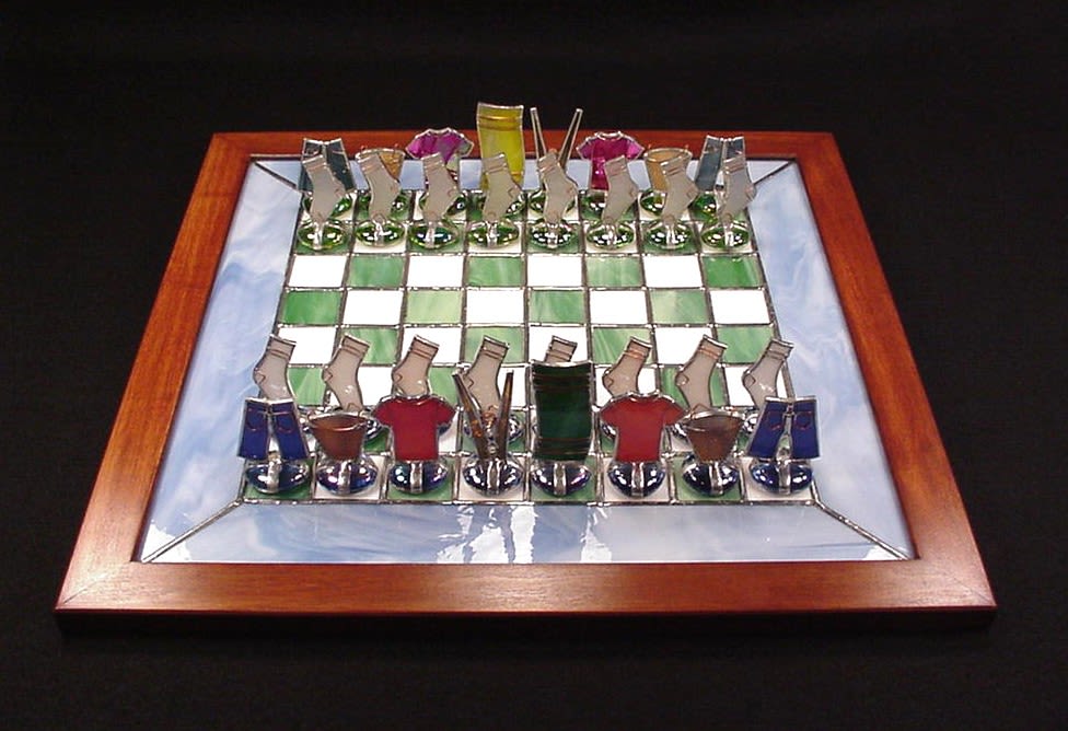 #A123 - Laundry Day Chess Set - A luminous stained-glass chessboard makes for an unforgettable gift that feels handcrafted, playful, and perfect as a decorative and usable art piece, ideal for Mother's Day or a one-of-a-kind present for Father's Day, an anniversary, or a birthday. This unique game board features green-and-white checkered stained glass board set inside a warm wooden frame, surrounded by a cloudy blue glass band that catches light and lends a serene, artisanal glow. The whimsical chess pieces - bright glass forms shaped like shirts, pants, and socks with polished metal bases - give the piece personality so the recipient will smile every time they see it.   The glasses in greens, whites, blues, and mixed hues accentuate the board's colors and handcrafted feel, creating a balanced display that works as a centerpiece or keepsake accent. It's an ideal choice for occasions from graduation and retirement to get well, new baby celebrations, thank you’s, or "just because" moments when you want someone to feel genuinely seen and delighted. Sized to sit on a coffee table or mantel, this one-of-a-kind chess set brings both visual texture and playful charm to holiday and everyday gifting moments.  Shipping available - select Delivery, then message us to ship.