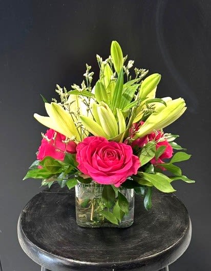 #A129 - Cube of Sunshine - Vivid hot pink roses and pale yellow lily buds make an energetic, cheerful statement in this compact flower arrangement - perfect as a birthday bouquet, Mother's Day gift, or a bright "thinking of you" surprise. The combination reads as both romantic and joyful, a lively way to say congratulations, thank you, or "get well soon" to the person you want to uplift. The flowers are tightly gathered with small white filler blooms for soft texture and contrast, arranged low and wide in a clear glass cube vase that shows the clean lines of the stems for a modern, approachable look.   The cube's compact dimensions make it ideal for tabletops, desks, and bedside stands; the bold pink-and-yellow palette also translates beautifully for spring and summer events or as a cheerful accent during autumn and holiday gatherings.  This arrangement suits a wide range of occasions-anniversary, graduation, housewarming, new baby, or simply "just because"-and the sturdy glass cube keeps it easy to display. For best longevity, change the water every other day and trim stems slightly when refreshing; ordering same-day or scheduled delivery ensures the recipient receives the freshest possible presentation.