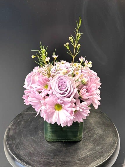 #A037 - Purple Passion - A compact dome of lavender roses anchors this “Purple Passion” flower arrangement, the gentle hue creating an immediate sense of calm and affection perfect for a birthday bouquet, anniversary roses, or a Mother's Day gift. Soft lavender daisy-style chrysanthemums form a ring around the roses, while tiny white filler blooms and slender buds lift the composition with an airy, delicate texture-an arrangement that makes the person you're thinking of feel seen and comforted. Arranged low in a green-lined square glass vase, the structured contemporary base contrasts the soft pastels for a modern, polished look that suits housewarming, graduation, get well, or just because moments.   The mix of daisies, roses, chrysanthemums and small white accents delivers layered color-lavender, pink, green and white-without being fussy, and the compact size fits easily on bedside tables, desks, or dining tables. Give this as a thoughtful thank-you, thinking-of-you, new baby, retirement, or romantic gesture; it also reads beautifully for holidays, Father's Day when you want something gentle and unexpected, or to say I'm sorry. Care is simple: keep water fresh, trim stems if needed, and display out of direct heat to extend vase life so she (or he) can enjoy the soft scent and serene palette for days.