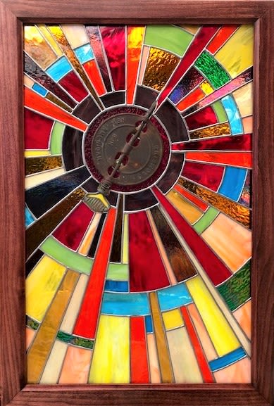 #A038 - Sun with Damper - Stained Glass Window - A radiant stained glass wall art piece that reads like a sunburst in color-this framed stained-glass panel is an artful gift for celebrations, housewarmings, or to brighten an everyday living space. The arrangement-like composition centers on an antique woodstove damper, with long, narrow glass "rays" fanning outward in vivid reds, yellows, oranges, blues and touches of green and cream. The result is an energetic, uplifting focal point that greets light with a lively, translucent glow. The piece pairs richly textured, translucent glass with a warm-toned wooden frame that grounds the vivid interior composition; the contrast between the smooth metal dial and the hand-cut glass adds tactile interest. Colors shift across the panel as light changes, making it a versatile accent for living rooms, foyers, or sunlit nooks.   Sized to hang on a wall as a single statement piece, it arrives ready to display and instantly transforms a room with color and motion. Give it to someone who deserves to feel seen-this is the kind of gift that reads as joyful and deliberate, perfect for birthdays, congratulations, or a thoughtful housewarming. Care is simple: hang where it can catch natural or artificial light and dust the frame and glass gently to preserve the luminous effect.