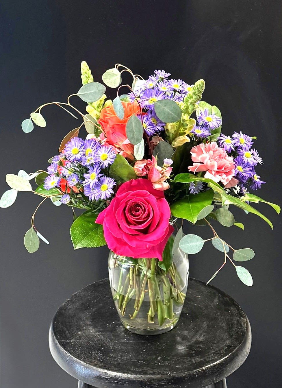 #A039 - Creating Sweet Memories - A vivid hot pink rose anchors this mixed flower arrangement, giving it a joyful, celebratory focal point perfect for a birthday bouquet, Mother's Day gift, or a "just because" surprise. The lively mix of pink, purple, white, yellow, orange, and peach reads like a sunlit garden - a look that also suits congratulations for graduation, a cheerful get-well message, or a housewarming present. The arrangement combines roses, carnations, chrysanthemums, asters, and snapdragons arranged in a clear glass vase. Soft peach and coral carnations and other accent blooms nestle around the focal rose while sprays of small purple asters with bright yellow centers add playful contrast. Taller snapdragon stems create vertical interest and the round, airy eucalyptus arcs outward for a loose, garden-inspired silhouette that feels fresh and natural.   This bouquet sends warmth and uplift to the person you're thinking of - whether you're marking an anniversary, saying thank you, welcoming a new baby, or brightening someone's winter day. Arrives ready-to-display in its glass vase; light daily watering and trimming stems every few days will keep blooms looking their best.