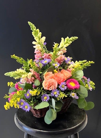 #A049 - Thinking of You - Peach roses clustered at the heart of this woven basket arrangement create a warm, inviting focal point-the perfect “thinking of you” gift to brighten someone's day. This flower arrangement is ideal for Mother's Day, birthdays, anniversaries, or “just because” blends garden-fresh charm with a cheerful palette so the person you're sending it to feels genuinely seen and uplifted. Soft pink and pale white snapdragons rise above the composition for graceful height, while purple aster-like blooms and bright yellow filler flowers weave texture and contrast throughout. Daisies, carnations, chrysanthemums, stock, sunflowers and a few snapdragon spikes fill the low, densely packed basket for a relaxed, abundant look that reads both casual and carefully arranged.   Give this as a get well, congratulations, new baby, retirement, or thank-you surprise-the combination of colors and mixed blooms reads joyful and reassuring across spring and summer occasions. The woven basket arrives ready to display; for best longevity, place in fresh water daily, and keep away from direct heat to help the recipient enjoy the flowers longer.