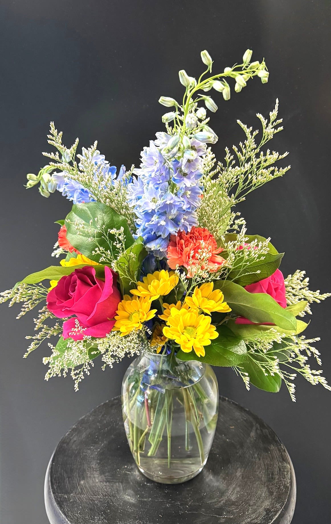 #A057 - Color Chaos - A joyful riot of color - tall blue delphinium stems soar above vivid pink roses, sunny yellow daisies, and cheerful orange carnations to create a bright flower arrangement perfect for birthdays, Mother's Day, or a cheerful Easter centerpiece. This flower arrangement reads as energetic and uplifting, the kind of bouquet that makes the person you're thinking of feel truly celebrated and noticed.  Crafted in a rounded clear glass vase with a neat blue ribbon, the mix also includes chrysanthemums, fine white filler clusters, and broad green foliage for contrast and a light, garden-style silhouette. The arrangement's medium height and water-filled vase make it an easy desk or dining-table statement; sizes are balanced so it works for graduations, get-well wishes, housewarmings, anniversaries, and "just because" moments.  Send this when you want to brighten someone's day-whether it's a thank-you for a thoughtful favor, congratulations on a retirement, or a loving surprise for a new baby. Light care: keep vase topped with fresh water and out of direct heat to prolong bloom life.