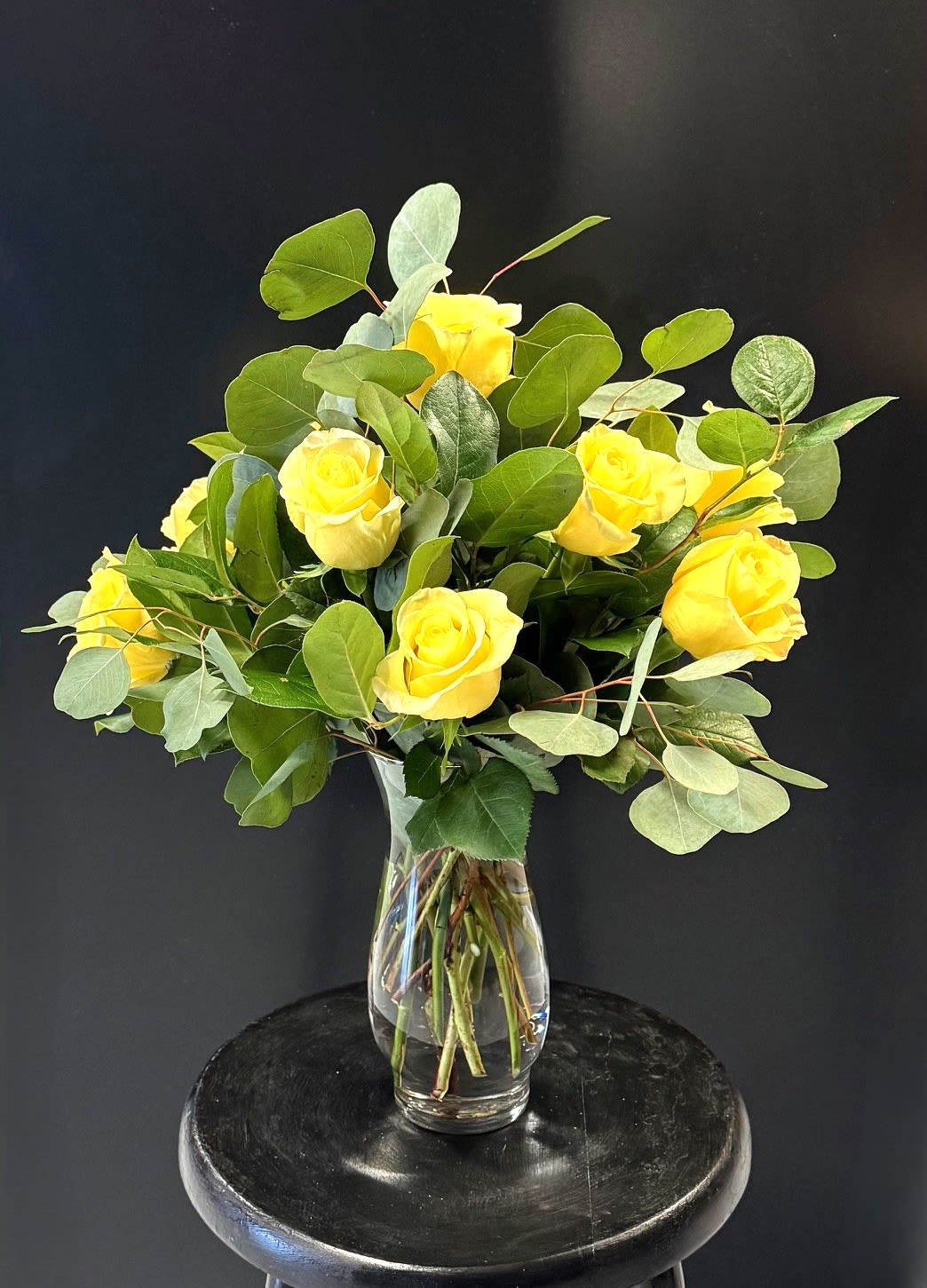 #A064 - Blossoming Friendship - Sunlit yellow roses take center stage in this cheerful flower arrangement, a bright and optimistic choice for a birthday bouquet, Mother's Day gift, or a heartfelt "thinking of you" surprise. The arrangement reads as both modern and approachable - perfect for Father's Day, anniversaries, housewarmings, graduations, or simply to lift someone's spirits when they need a pick-me-up. A dozen lively yellow roses are nestled into abundant broad, rounded deep-green foliage for a full, airy silhouette that feels generous without being fussy. The blooms are displayed in a simple clear glass vase that shows off the stems for a clean, classic look; in the pictured styling the vase sits on a round black stool, giving the composition a contemporary edge that complements autumn and winter settings just as well as spring and summer.   Choose this arrangement to celebrate new babies, retirements, weddings, or to say thank you, I'm sorry, congratulations, or get well soon-the warm yellow tones make the recipient feel genuinely seen and uplifted. Sized to fill a room with color and texture, the vase makes care easy: refresh the water every few days and trim stems at an angle for longest life. Order with a note to tailor the message for any occasion, from holidays to everyday acts of kindness.