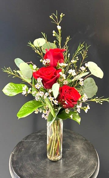#A072 - 3-Of-A-Kind - Three vivid red roses stand upright in a slim clear glass cylinder vase, making this flower arrangement an elegant and unmistakable symbol of love and celebration. As a birthday bouquet, anniversary roses, or a simple "thinking of you" gift, the vertical, airy design keeps the focus on the three blooms so the person you're honoring will feel cherished. This arrangement also suits Mother's Day, Valentine's Day, or a romantic surprise without feeling overdone. Delicate white filler flowers weave between the roses, adding soft contrast and a light, textured feel against the rich red and fresh green stems.   The minimalist composition and clear glass vase highlight the natural lines of each stem and keep the display compact enough for a desk, bedside table, or mantel. Ideal for occasions from Graduation and Get Well to Retirement, Housewarming, or "I'm Sorry," it reads as thoughtful whether given for a major milestone or just because. The slim cylinder vase and upright styling create a neat, low-maintenance arrangement.  Refresh the water every few days and recut stems after 4-5 days to extend vase life. Available year-round and seasonally fitting for Spring, Summer, Autumn, and Winter holidays, this trio-of-roses arrangement is the kind of floral gift that makes someone feel genuinely celebrated.
